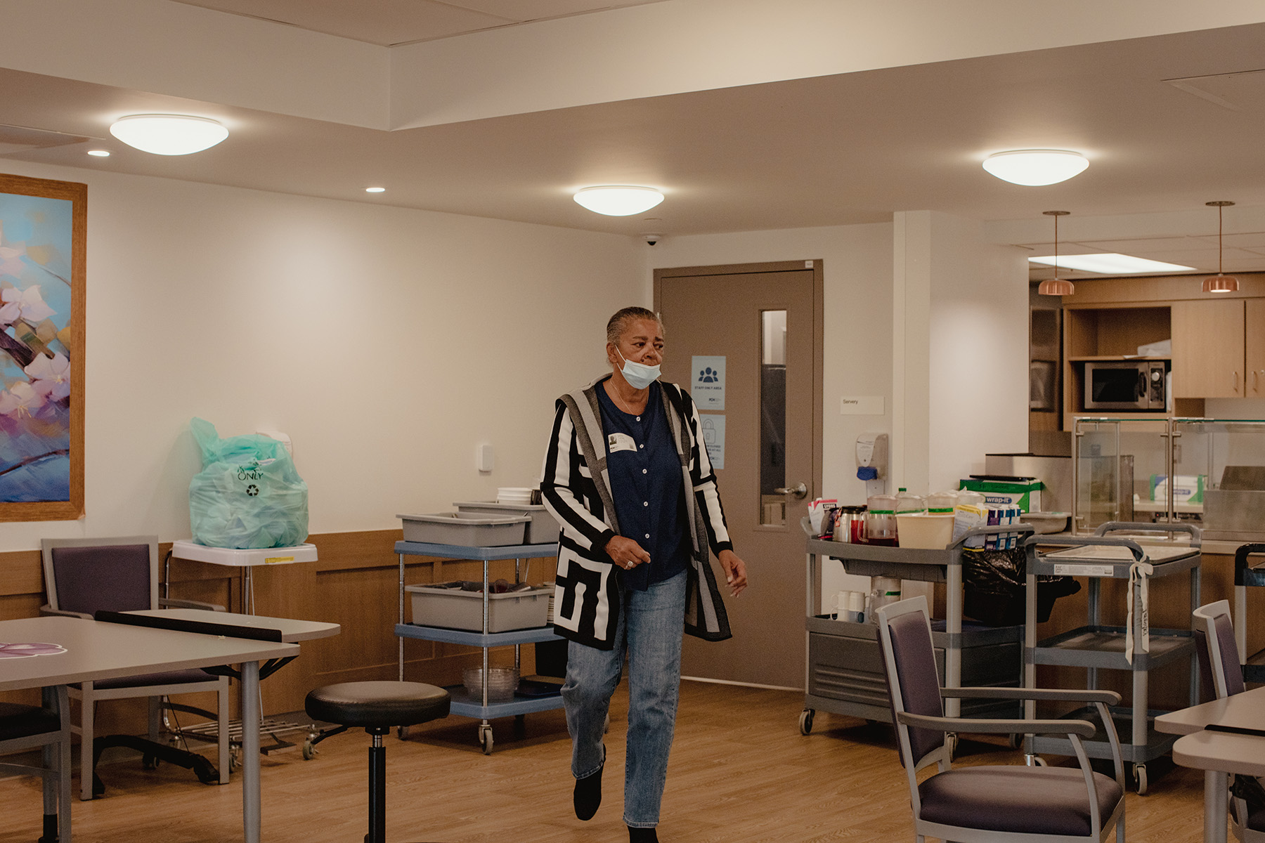 Left, Sandra Tierney helps cut food for a Wellbrook Place resident seated at her father’s table. Right, Sandra walks through the dining room after clearing dishes.